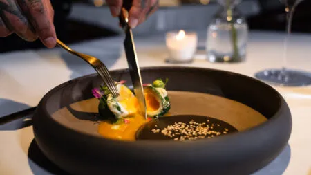 Chef using knife and fork to plate a gourmet dish with orange sauce, greens, edible flowers, and sesame seeds in a matte black bowl (candlelit table).