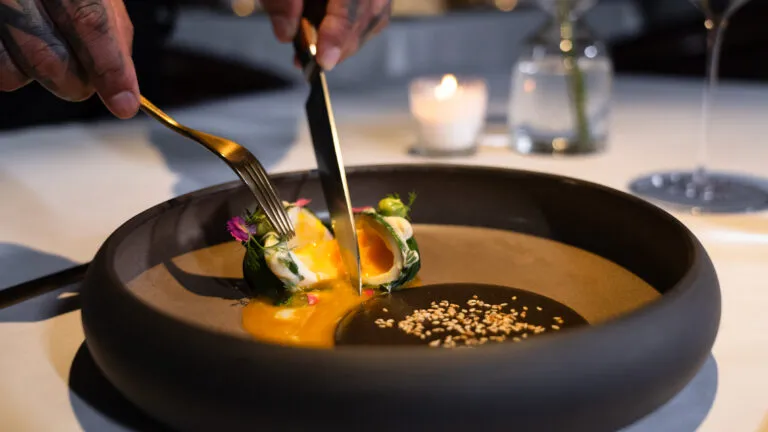 Chef using knife and fork to plate a gourmet dish with orange sauce, greens, edible flowers, and sesame seeds in a matte black bowl (candlelit table).