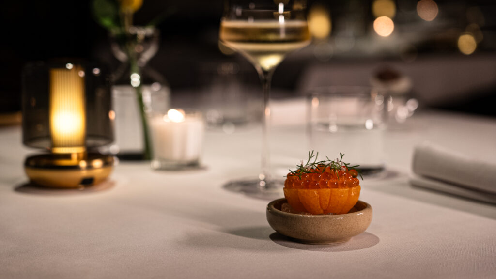 María Dolores by Edgar Núñez Haute Grandeur Global Awards 4 - Petróleo y Energía Close-up of a small orange cake topped with red roe and dill, plated on a ceramic dish in a fine-dining setting with blurred glasses in the background.