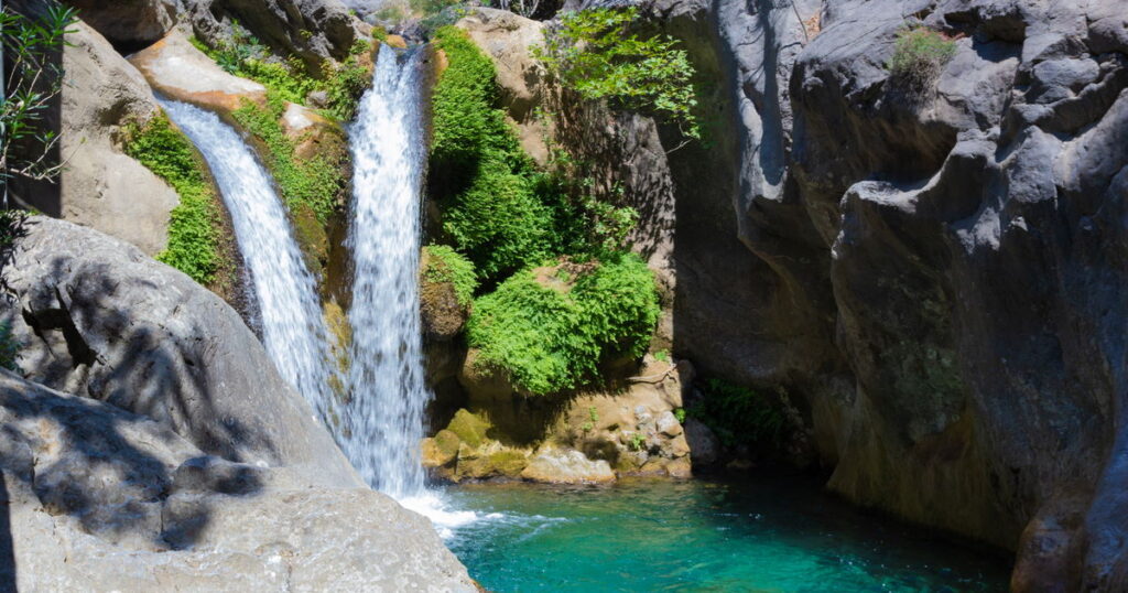 Sapadere-canyon-in-Antalya-in-Turkey - Petróleo y Energía Turquoise pool at the base of a two-tier waterfall cascading between rocky cliffs with green plants along the edges of a canyon.