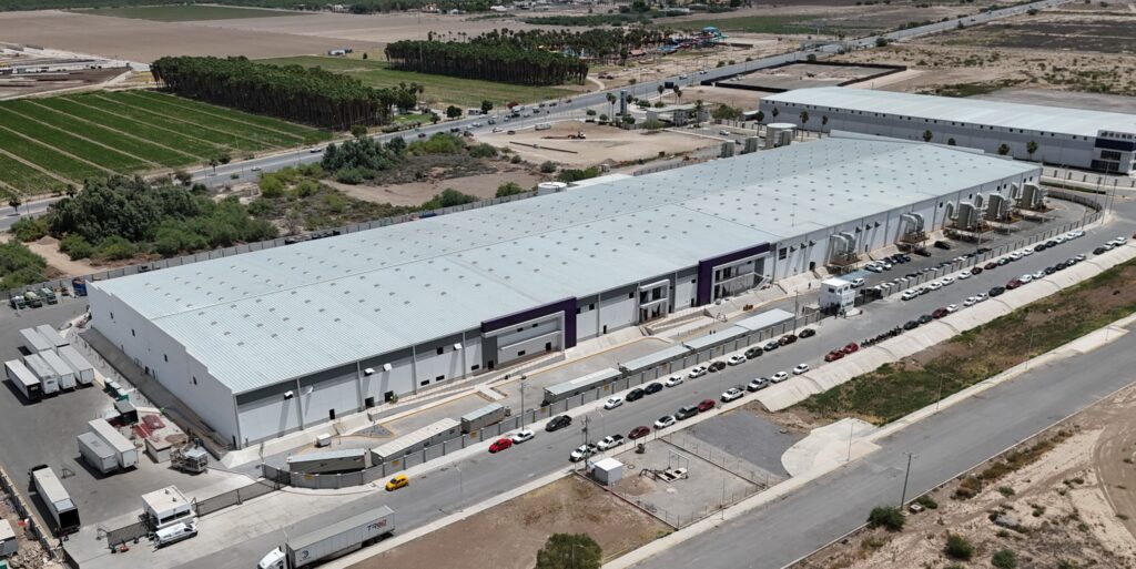 WhatsApp Image 2026-07-01 at 51449 PM - Petróleo y Energía Aerial view of a large industrial complex with long white-roof warehouses, loading docks, and a row of parked cars and trucks along the street beneath a clear sky.