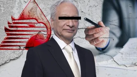Man in a suit with eyes blacked out stands beside a large red graffiti emblem on a cracked wall; a hand holding a pen approaches as if about to sign.