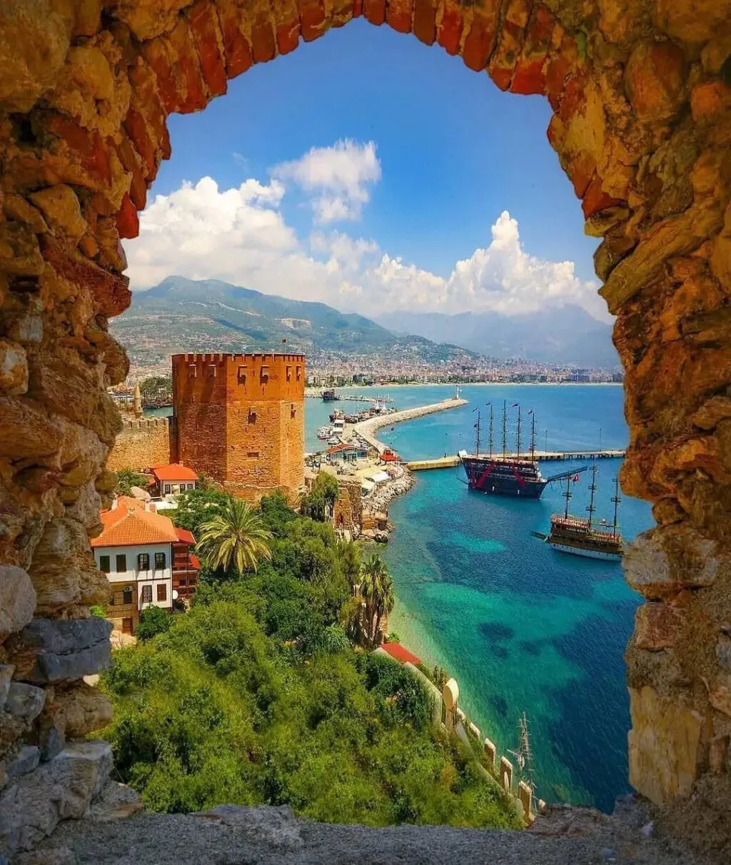 View of a historic red-brick fort by a turquoise bay, framed by a stone arch with boats in the harbor and distant mountains.