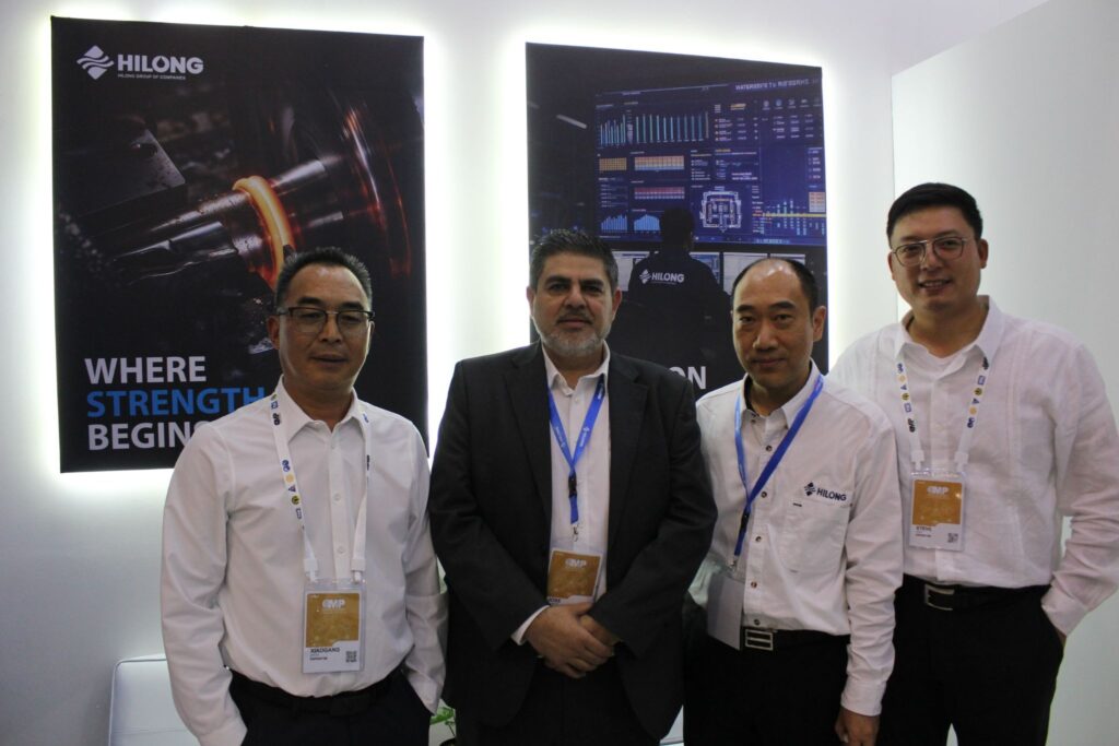 image - Petróleo y Energía Group of four men in white shirts with lanyard badges posing in front of Hilong banners at a booth.