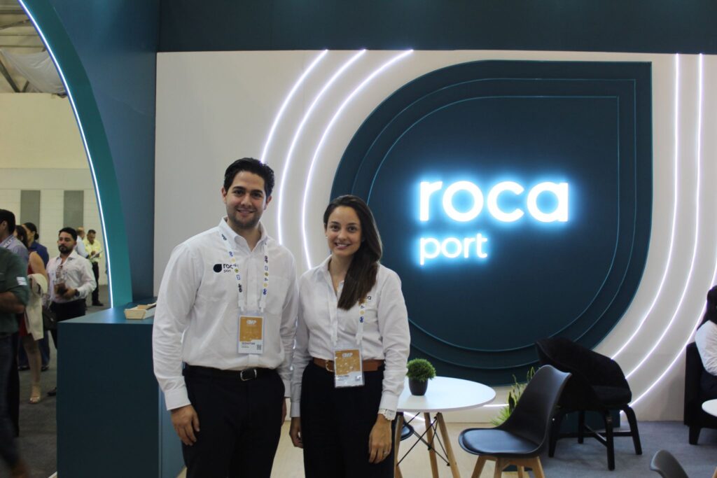 image - Petróleo y Energía Two exhibition staff in white shirts pose at a ROCa Port booth with teal circular arches and neon lighting behind them.