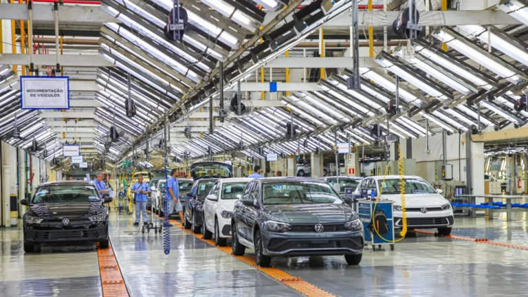 Automotive assembly plant with multiple cars on a moving bright orange conveyor in a spacious, brightly lit aisle; workers in blue uniforms inspect vehicles under overhead machinery.